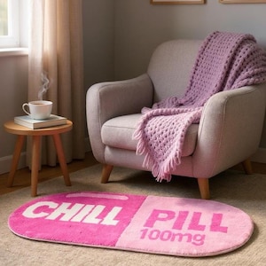 May include: A pink and light pink rug shaped like a pill with the words "CHILL PILL" and "100mg" printed on it. The rug is in front of a gray armchair with a purple blanket. A small wooden side table holds a cup of coffee.