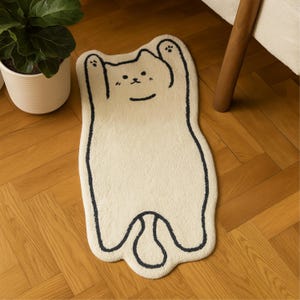 May include: Cream-coloured cat-shaped rug with a black outline and facial features. The rug has a playful design, with the cat's paws raised. The rug is placed on a wooden floor, near a potted plant and a chair leg.