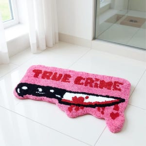 Puede incluir: Alfombra de baño rectangular rosa con las palabras "TRUE CRIME" en rojo. La alfombra presenta un diseño de cuchillo en blanco y negro con salpicaduras de sangre roja y detalles de corazón.