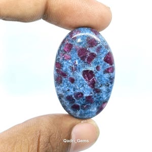 Op de afbeelding: Een ovaalvormige edelsteen met een blauwe achtergrond en ingebedde rode kristallen. De steen wordt tussen twee vingers gehouden tegen een witte achtergrond. De tekst "Qadri_Gems" is zichtbaar onderaan.