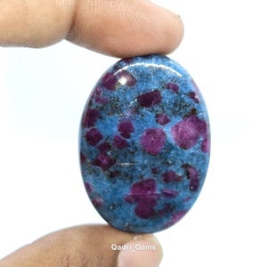 Könnte beinhalten: Ovaler Edelstein mit blauem Grund und tiefroten Einschlüssen. Der polierte Stein hat eine glatte, glänzende Oberfläche. Der Edelstein wird zwischen Fingern vor weißem Hintergrund gehalten. Der Text "Qadri_Gems" ist unten sichtbar.