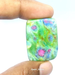 May include: A rectangular gemstone with a vibrant green base, speckled with pink, blue, and yellow hues. The stone is held between two fingers. The text "Qadri_Gems" is visible at the bottom.