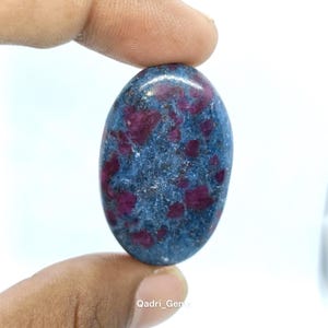 May include: An oval gemstone featuring a blue matrix with striking red inclusions. The polished stone is held, revealing its smooth surface and natural colour variations. A unique piece for jewellery or collection.