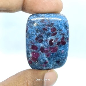 May include: A rectangular gemstone with a blue base color and scattered red inclusions. The stone is held between fingers, with the text "Qadri_Gems" visible at the bottom.