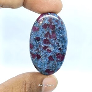 May include: Oval-shaped gemstone with a blue-grey matrix and deep red inclusions. The polished stone is held between fingers, showcasing its natural patterns and textures. The gem is a unique piece of jewelry.