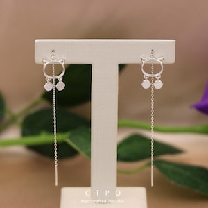 Könnte beinhalten: Ein Paar silberne Ohrringe in Katzenform. Jeder Ohrring hat einen Katzenumriss, zwei baumelnde Anhänger und eine lange Kette. Die Ohrringe werden auf einem neutralen Ständer präsentiert, mit dem Text "CTPO Handcrafted Jeweller" unten.