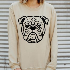 May include: A beige sweatshirt featuring a large black graphic of a bulldog's face. The bulldog's face is centered on the front of the sweatshirt. The sweatshirt has a crew neck and long sleeves.