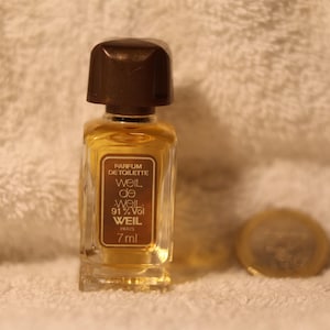 Può includere: Un piccolo flacone di profumo con un tappo marrone scuro e una forma rettangolare. Il flacone contiene un liquido dorato e ha un'etichetta con la scritta "Parfum de Toilette Weil Paris". Il flacone è da 7 ml.
