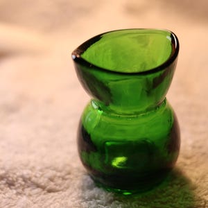 Op de afbeelding: Een kleine, groene glazen vaas met een unieke zandloper vorm. De vaas heeft een brede, open bovenkant en een afgeronde basis. Het glas is een levendig, transparant groen en de vaas staat op een getextureerd oppervlak.