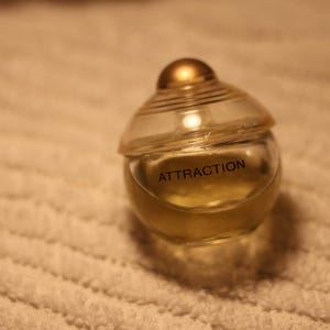 Könnte beinhalten: Ein kleiner, klarer Glasparfümflakon mit einer goldfarbenen Flüssigkeit im Inneren. Die Flasche hat eine abgerundete Form mit einem goldfarbenen Verschluss und dem Wort "ATTRACTION" auf der Vorderseite. Die Flasche steht auf einer weißen, strukturierten Oberfläche.