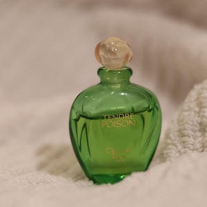 Könnte beinhalten: Eine grüne Parfümflasche mit einem klaren, abgerundeten Verschluss. Die Flasche hat eine geschwungene Form und goldfarbene Schriftzüge mit der Aufschrift "Tendre Poison" und "Christian Dior". Die Flasche steht auf einer weißen, strukturierten Oberfläche.