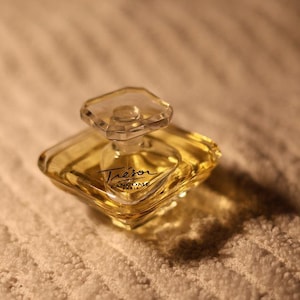 Può includere: Un flacone di profumo trasparente a forma di diamante con un tappo quadrato trasparente. Il flacone contiene un liquido giallo dorato e presenta la scritta "Trésor" in corsivo. Il flacone di profumo è su una superficie bianca strutturata.