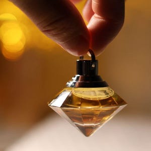 Könnte beinhalten: Ein rautenförmiger Parfümflakon mit dunklem Verschluss und kleinem Ring. Die Flasche ist mit einer goldenen Flüssigkeit gefüllt. Der Hintergrund ist verschwommen mit warmen, gelben Tönen.