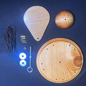 May include: A wooden craft kit with various components, including a round base, a teardrop-shaped piece, a sphere, and hardware. The kit also includes a bundle of black cord and several screws and washers. The items are arranged on a dark blue surface.