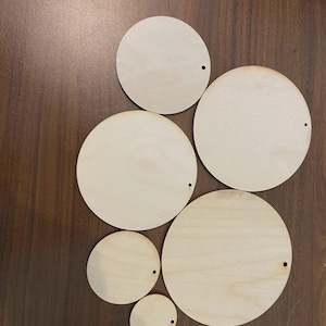 May include: A collection of unfinished, light-colored wooden circles of varying sizes, each with a small hole near the edge. The circles are arranged on a dark brown wooden surface. These wooden circles are suitable for crafts and DIY projects.