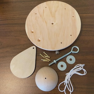 May include: A wooden craft kit with a round base, teardrop-shaped piece, and a spherical component. Includes screws, a bolt with an eye, washers, and white cord. The components are arranged on a brown wooden surface.