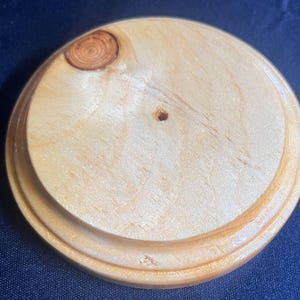 May include: A round, unfinished wooden base with a light brown color. The base has a raised edge and a small hole in the center. A knot is visible on the top surface. The wood grain is visible.