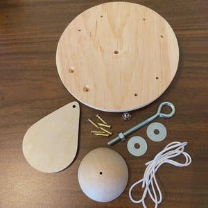 May include: A wooden craft kit with a round base, teardrop shape, and spherical component. Includes screws, a bolt with an eye, washers, a nut, and white cord. The components are made of unfinished wood.