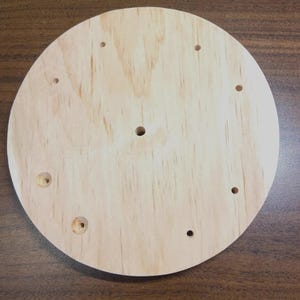 May include: A round, unfinished wooden disc with multiple pre-drilled holes. The light-colored wood grain is visible, and the disc appears ready for assembly or further customization. The holes are of varying sizes.