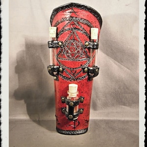 Puede incluir: Protección de brazo de cuero rojo con detalles negros y un diseño geométrico. Tiene tres pequeños viales de vidrio y un vial más grande unido a él.