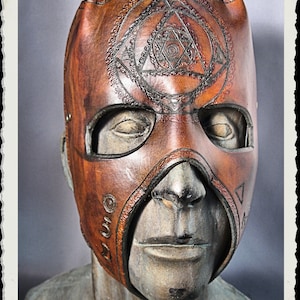 May include: A brown leather mask with intricate geometric designs carved into the surface. The mask has a pointed nose and eye holes, and is worn on a mannequin's head.