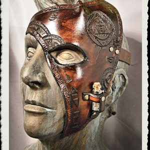 May include: A brown leather mask with intricate designs and a small vial attached to the side. The mask covers one eye and has a strap that goes around the head.