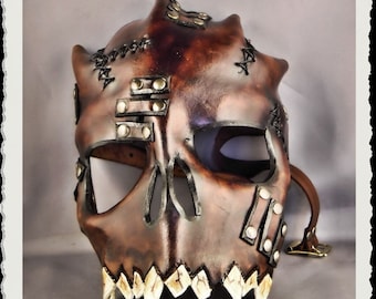 May include: A brown leather skull mask with stitched details, metal rivets, and a toothy grin. The mask has a strap for wearing.