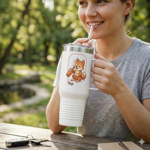 May include: White insulated tumbler with a handle and a lid. The tumbler features a cartoon fox illustration holding a bottle, with the word "SIP" below. The tumbler is being held by a person outdoors.