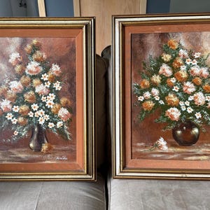May include: Two framed floral oil paintings. Each depicts a bouquet of flowers in a vase, with orange, white, and brown hues. The frames are gold and brown.