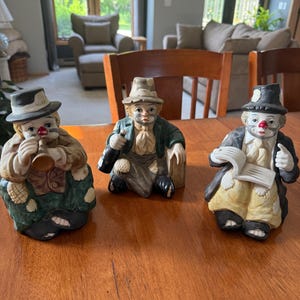 Puede incluir: Tres figuras de payasos de cerámica. Los payasos llevan sombreros y tienen narices rojas pintadas. Un payaso toca la trompeta, otro sostiene una botella y el tercero sostiene un pergamino. Las figuras están sobre una mesa de madera.