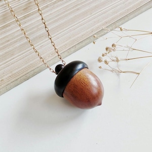 May include: A wooden acorn pendant necklace with a gold-colored chain. The acorn has a brown body and a black cap. The necklace is displayed on a white surface with a book in the background.