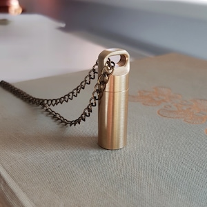 May include: A small, cylindrical brass pendant with a loop at the top, attached to a delicate chain. The pendant is a warm gold color and appears to be a container. The chain is a darker, antique brass color. The pendant rests on a light-colored, textured surface.