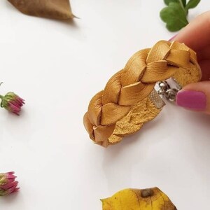 Könnte beinhalten: Ein geflochtenes Lederarmband in senfgelber Farbe. Das Armband hat einen silbernen Verschluss.