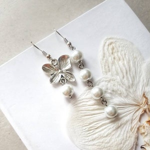 Puede incluir: Un par de pendientes de plata con un dije de flor y gotas de perlas blancas.