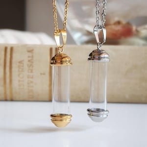 Puede incluir: Dos collares con viales de vidrio, uno con cadena y detalles dorados, el otro con cadena y detalles plateados. Ambos viales son cilíndricos y cuelgan de delicadas cadenas. El fondo incluye un libro borroso y una superficie blanca.