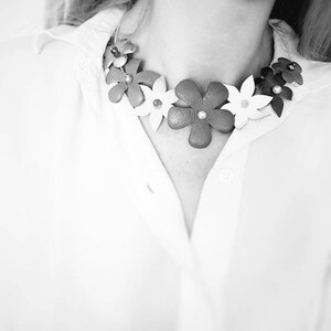 Peut inclure: Un chemisier blanc avec un collier de fleurs en cuir. Le collier est compos&eacute; de fleurs blanches, rouges et brunes.