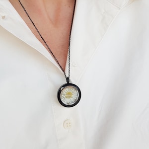 May include: A black chain necklace with a circular pendant. The pendant has a black frame and a clear center, showcasing a white and yellow flower. The necklace is worn over a white button-down shirt.
