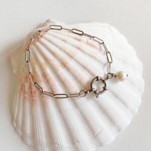 Könnte beinhalten: Ein silberfarbenes Kettengliederarmband mit einem großen Verschluss und einem einzelnen Perlenanhänger. Das Armband wird auf einer weißen Muschelschale präsentiert, wodurch eine Küstenästhetik entsteht. Die Glieder des Armbands sind rechteckig, und die Perle ist rund.