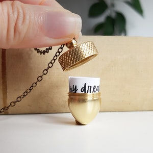 May include: A gold acorn necklace with a small, removable lid. The lid is open, revealing a small piece of paper with the words "my dreams" written on it.