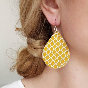 Könnte beinhalten: Ein Paar gelbe tropfenförmige Ohrringe mit einem weißen geometrischen Muster. Die Ohrringe sind aus Leder und haben silberne Haken.