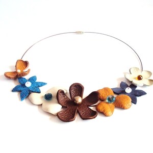 Peut inclure: Un collier en cuir avec un motif floral. Le collier pr&eacute;sente cinq fleurs dans des tons de brun, bleu, blanc et jaune. Chaque fleur a un petit cristal au centre.