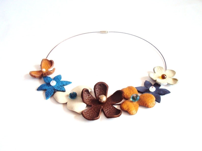 Peut inclure: Un collier en cuir avec un motif floral. Le collier pr&eacute;sente cinq fleurs dans des tons de brun, bleu, blanc et jaune. Chaque fleur a un petit cristal au centre.
