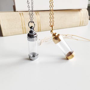 May include: Two glass vial necklaces with metal caps and chains. One necklace has a silver chain and cap, the other has a gold chain and cap. The vials are clear and cylindrical, designed to hold small items.