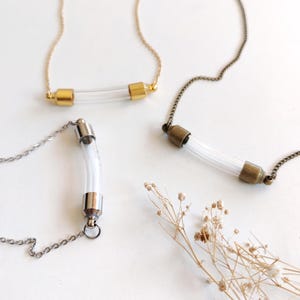 Puede incluir: Cuatro collares con colgantes de tubos de vidrio transparente. Dos collares tienen cadenas y detalles dorados, uno tiene una cadena plateada y el último una cadena de bronce. Los tubos de vidrio son cilíndricos y parecen vacíos. Una ramita de flores secas está en el fondo.