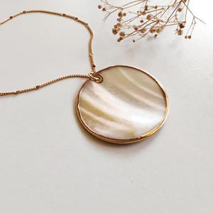 May include: A gold necklace with a large, circular pendant. The pendant has a pearlescent, iridescent surface within a gold frame. The delicate chain is also gold, with small beaded accents. A sprig of dried flowers is in the background.