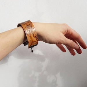 Puede incluir: Un brazalete de cuero marrón con un patrón floral y un dije de plata. El brazalete es ajustable y tiene un cierre de broche.