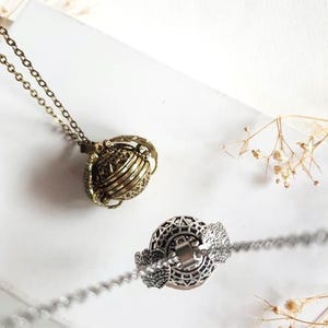 Könnte beinhalten: Zwei verzierte, kugelförmige Anhänger-Halsketten. Einer ist antikgoldfarben, der andere silberfarben. Beide haben aufwendige, durchbrochene Designs und hängen an zarten Ketten. Die Anhänger scheinen sich zu öffnen und Innenfächer freizugeben. Der Hintergrund ist weiß mit getrockneten floralen Akzenten.