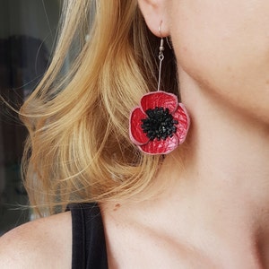 Peut inclure: Boucles d'oreilles en forme de coquelicot en cuir rouge avec un centre noir. Les boucles d'oreilles sont suspendues à un crochet argenté.