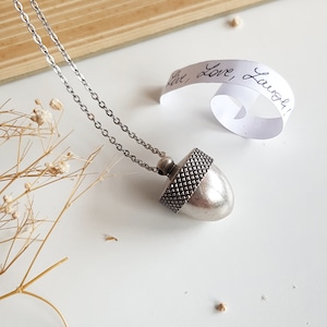May include: A silver acorn pendant necklace with a chain. The pendant has a textured top and a smooth bottom. The necklace is on a white surface with a small piece of paper that says "Live, Love, Laugh!"