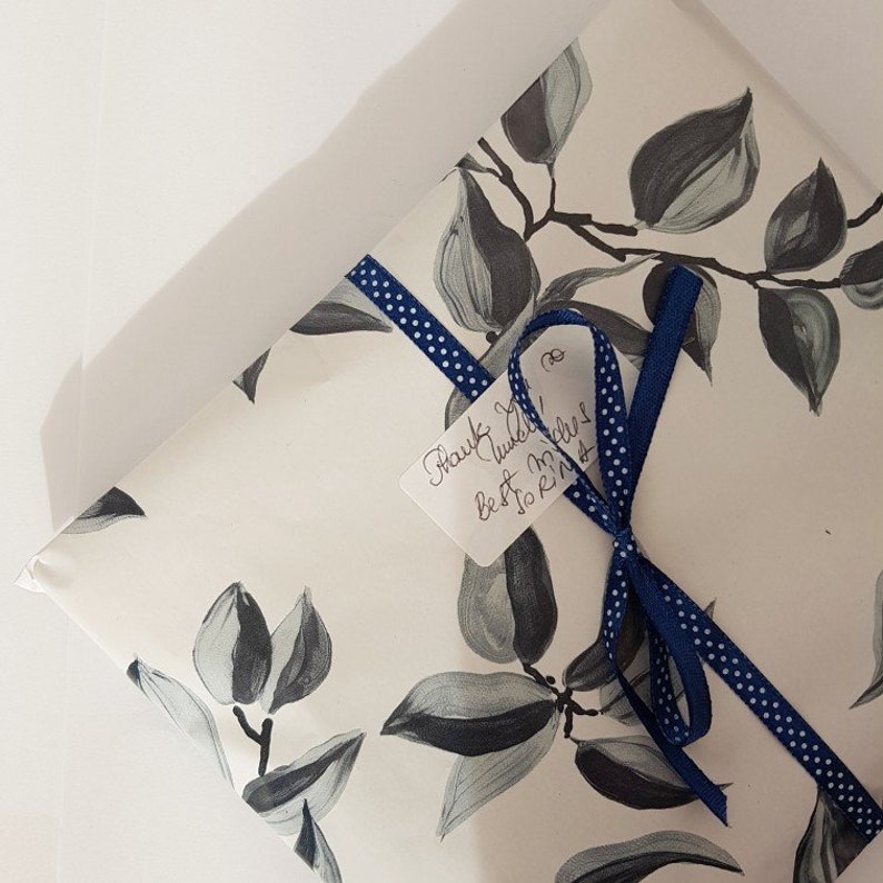 Peut inclure: Une bo&icirc;te cadeau blanche avec un ruban bleu et blanc et une note manuscrite qui dit "Merci ! Meilleurs v&oelig;ux & amour." La bo&icirc;te est emball&eacute;e dans du papier avec un motif de feuilles noires et blanches.